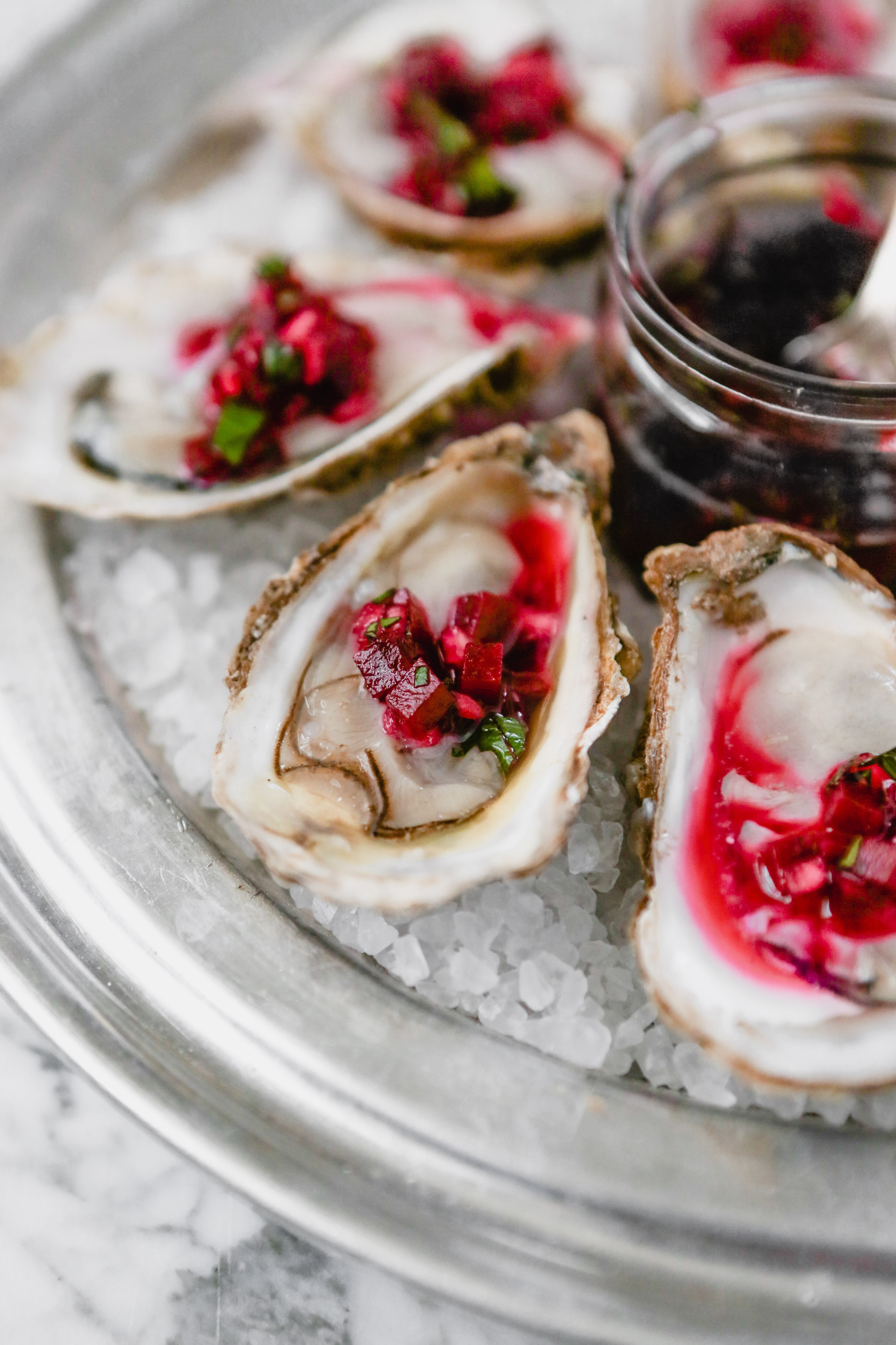 Photograph of oysters on the half shell set on rock salt in a metal pan on a marble surface, topped with a red mignonette sauce.
