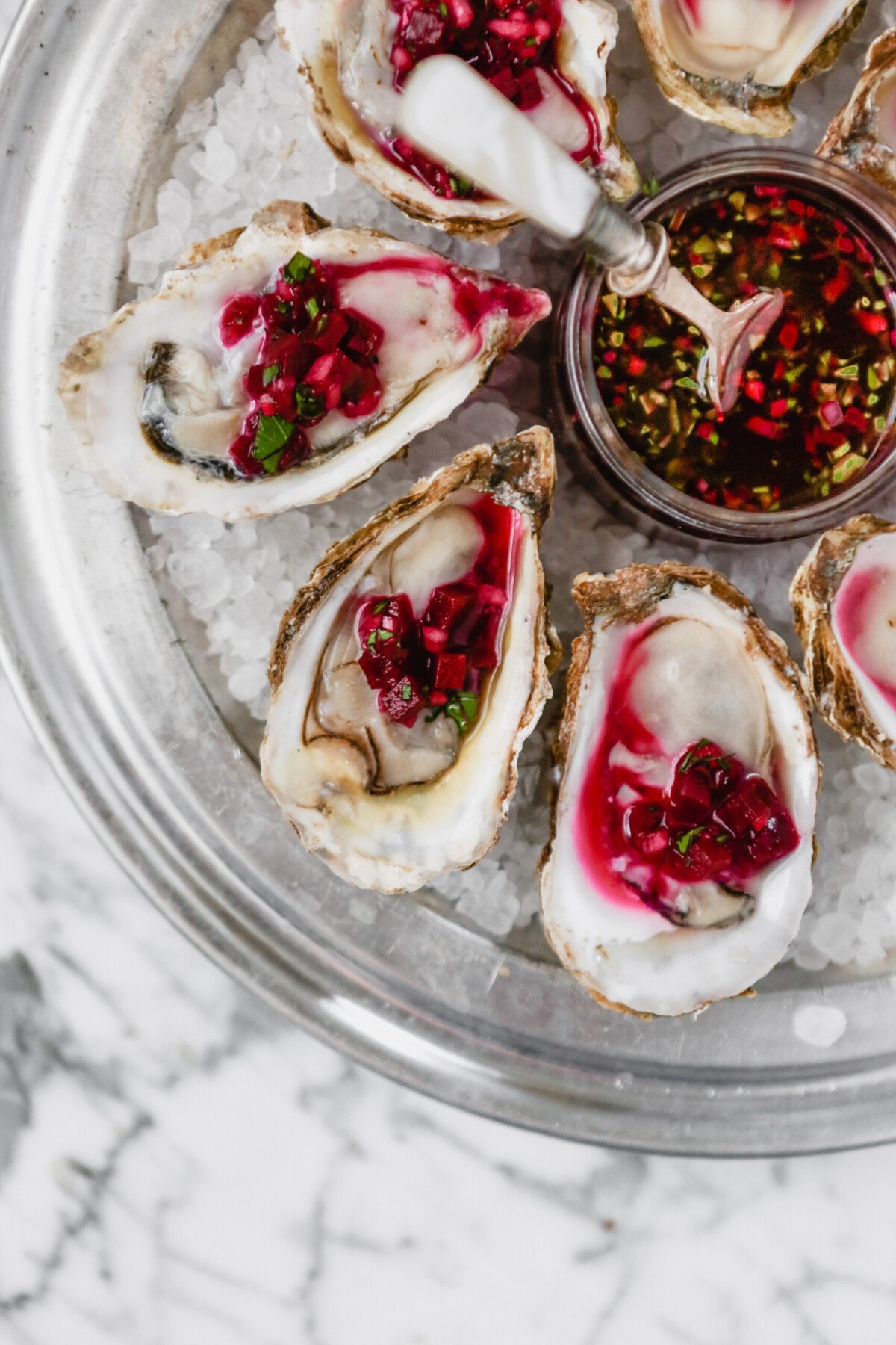 Photograph of oysters on the half shell set on rock salt in a metal pan on a marble surface, topped with a red mignonette sauce.