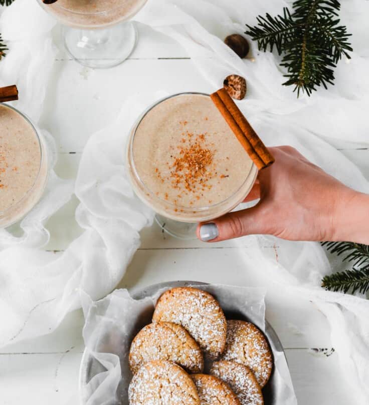 Photograph of homemade eggnog cookies and homemade vegan eggnog set on a white wooden table with evergreens