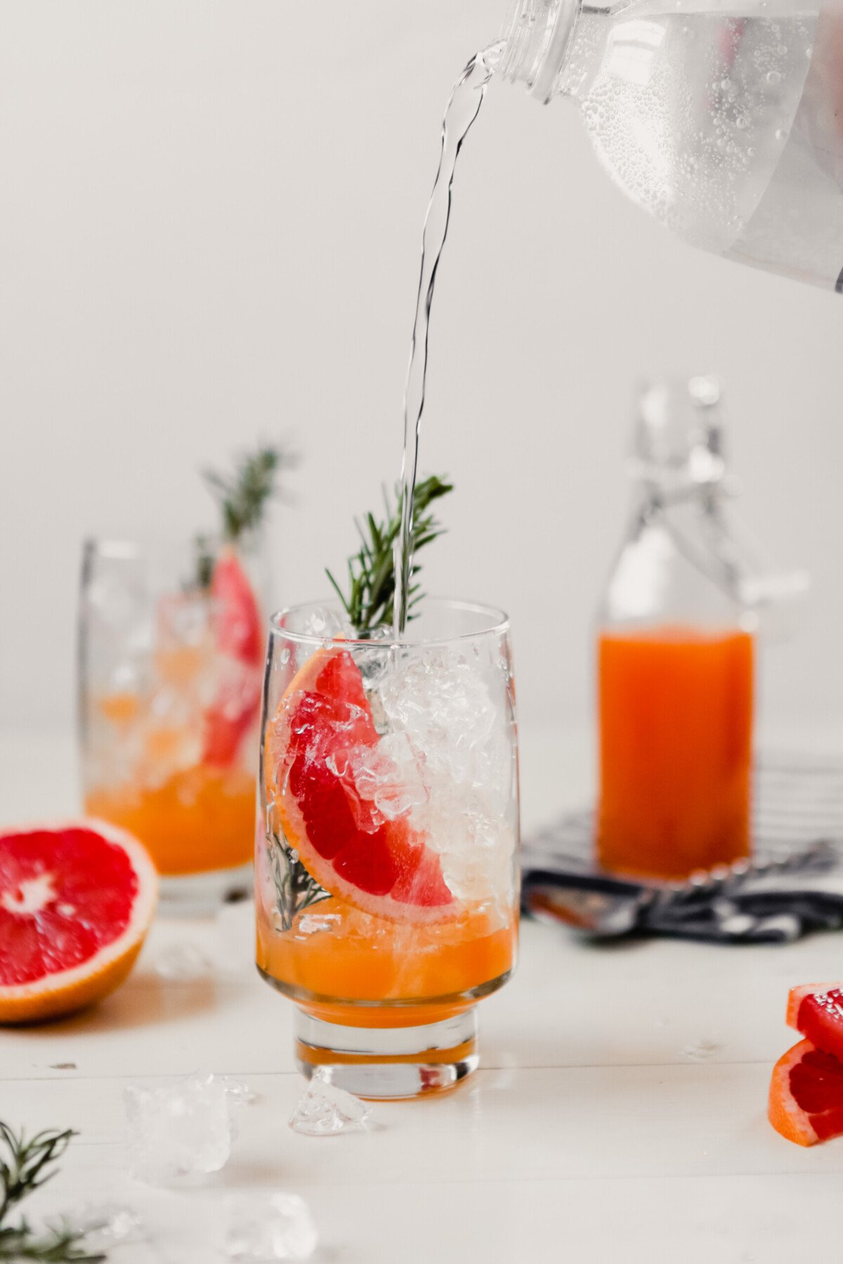 Photograph of club soda being pouring into .a glass with a grapefruit slice and sprig of rosemary to make a grapefruit soda.