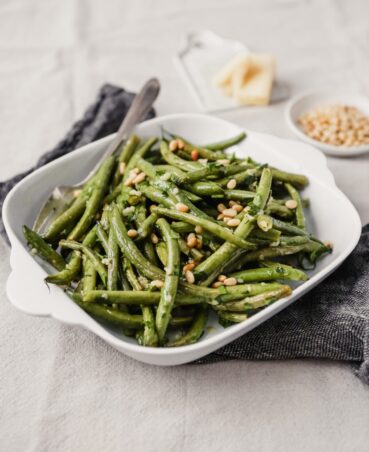 Roasted green beans with pine nuts in a square white serving dish. Parmesan and pine nuts in the background