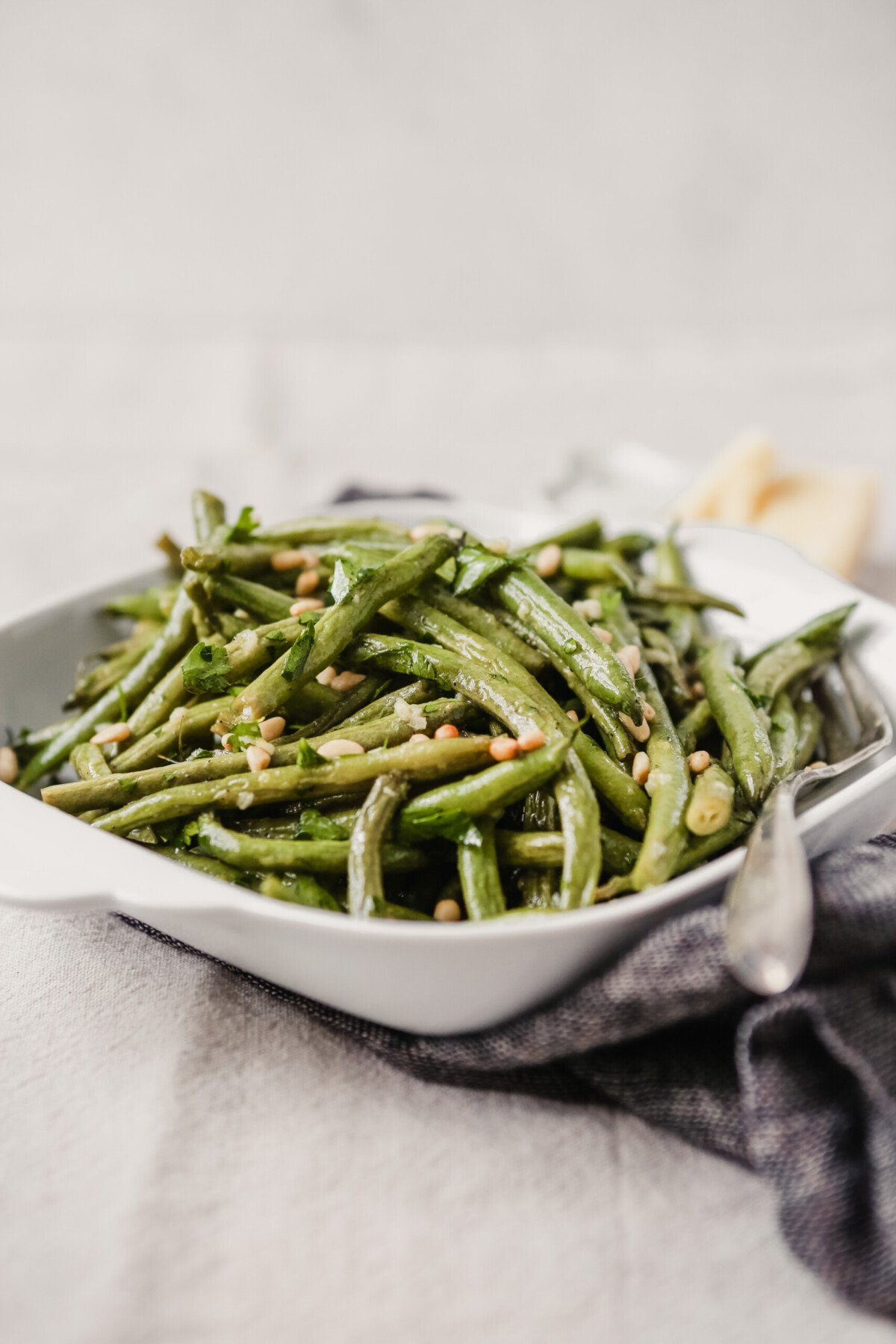 Roasted green beans with pine nuts in a square white serving dish. Parmesan and pine nuts in the background