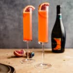 bright orange cocktail in two champagne flutes set on a brown table with a bottle of sparkling wine in the background