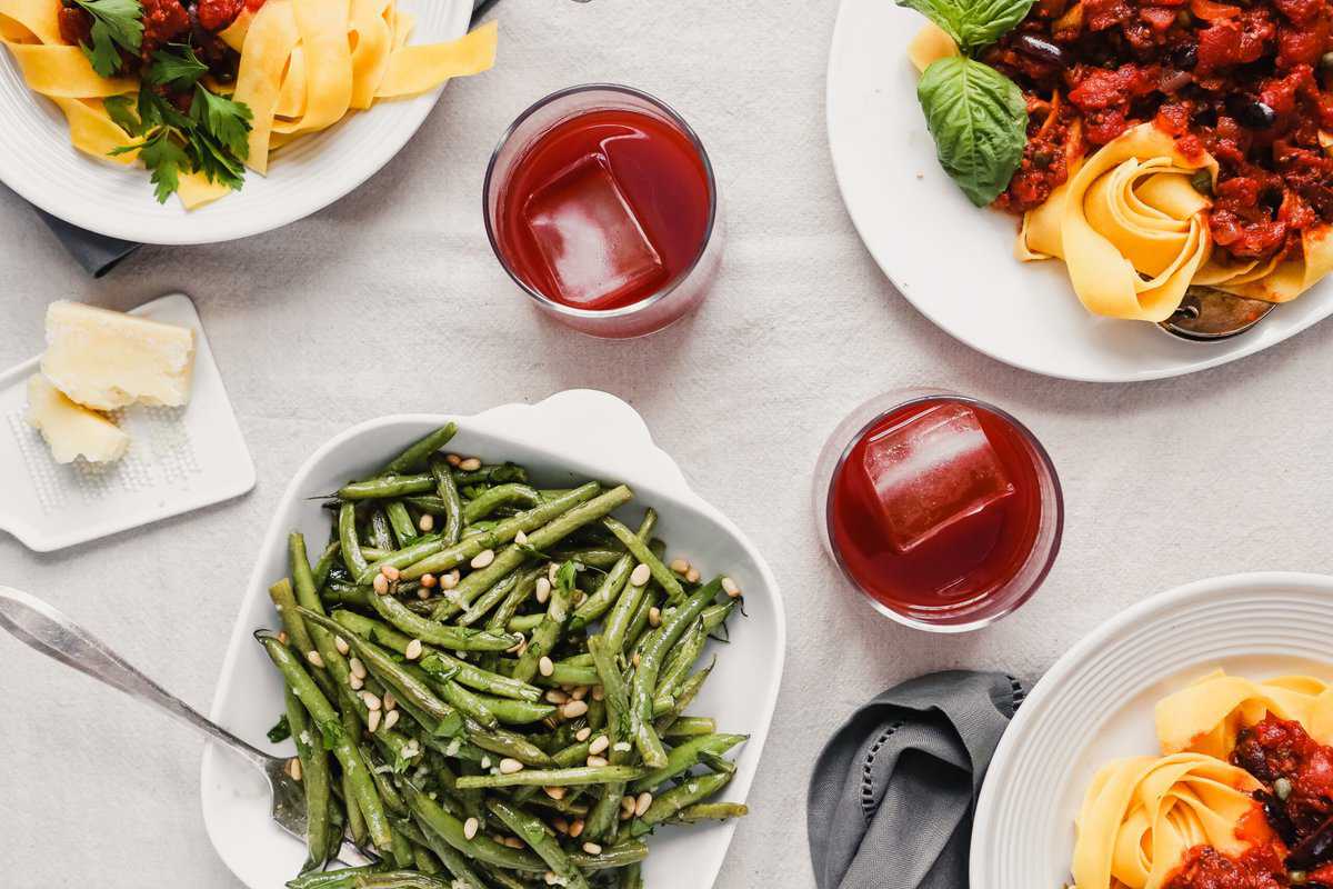 Photograph of an Italian dinner spread, green beans in a white bowl, glasses of Negroni and plates of pasta with red sauce