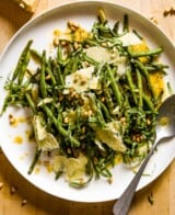 Roasted green beans, vinaigrette, cheese, pine nuts and basil on a large white serving plate with a silver brushed spoon.