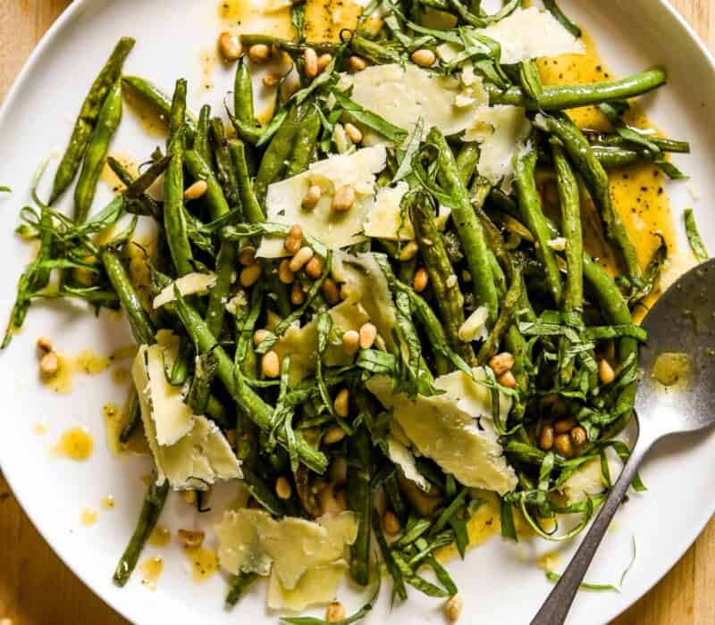 Roasted green beans, vinaigrette, cheese, pine nuts and basil on a large white serving plate with a silver brushed spoon.