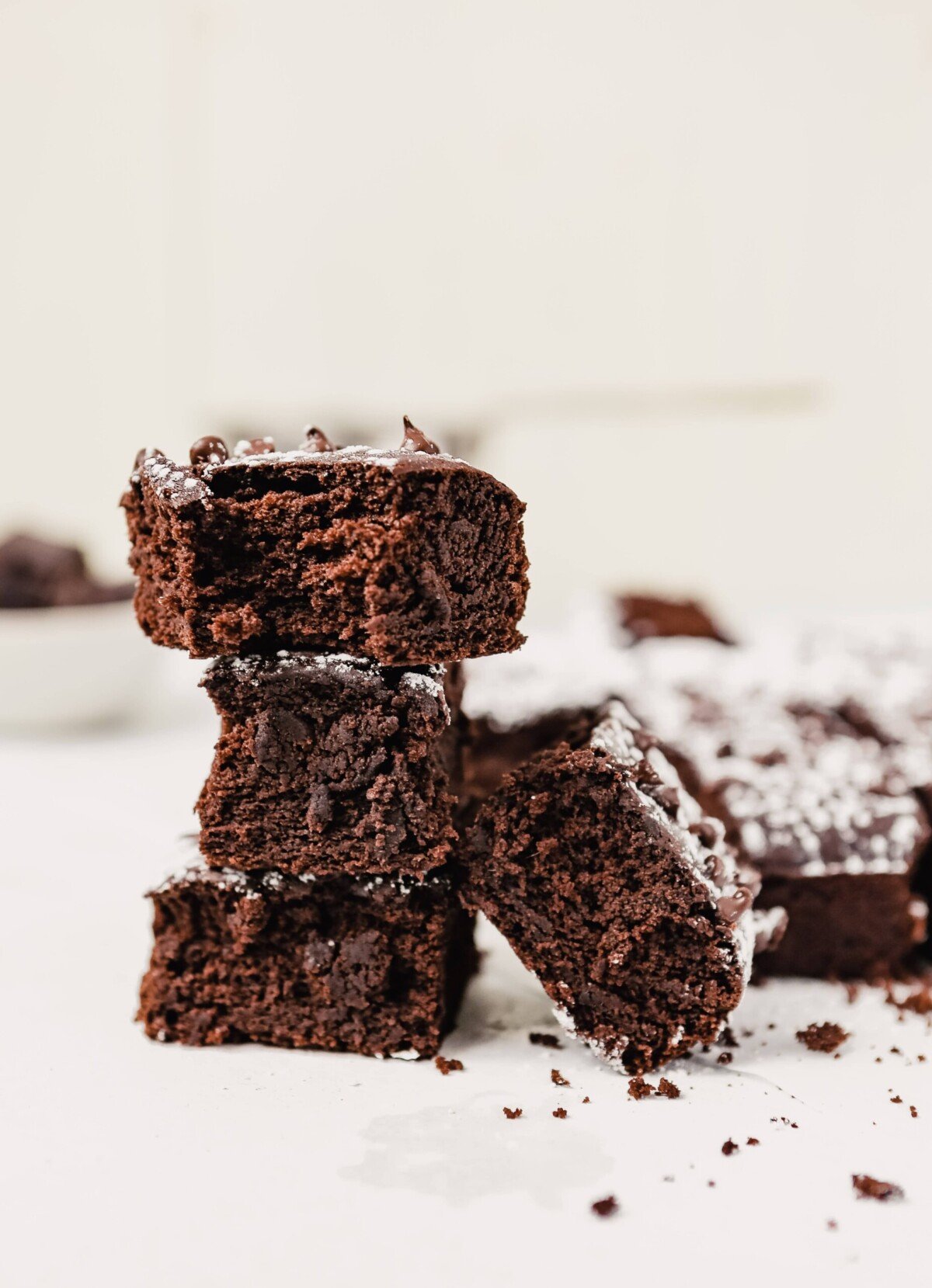 Photograph of a batch of brownies stacked on top of eachother on a white surface cut into squares and dusted with powdered sugar