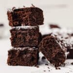 Photograph of a batch of brownies stacked on top of eachother on a white surface cut into squares and dusted with powdered sugar