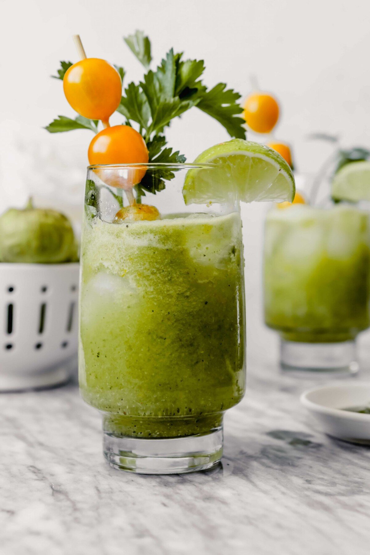 Photograph of green bloody mary in tall collins glass garnishes with yellow cherry tomatoes, herbs, lime wedge and apple slice.