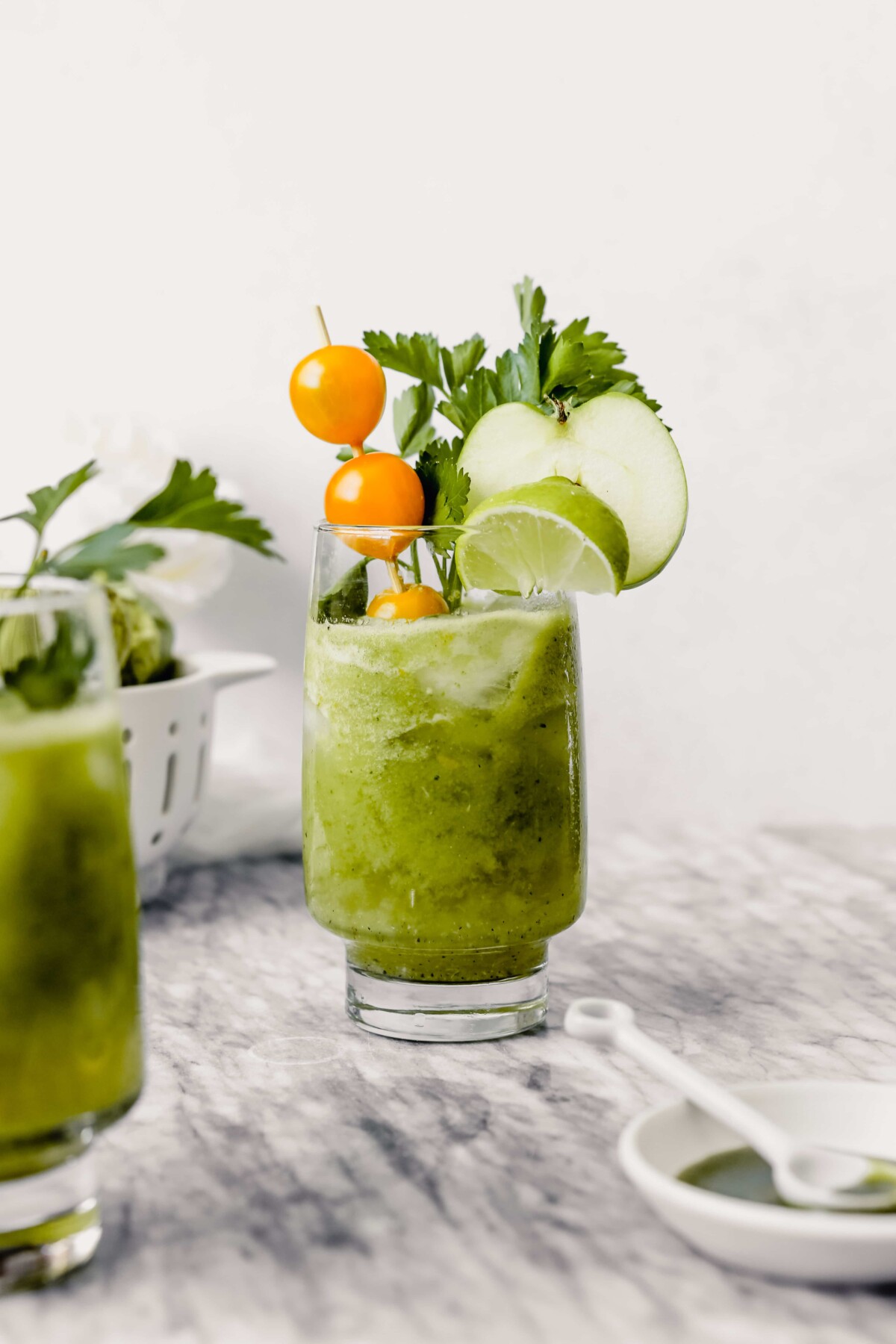 Photograph of green bloody mary in tall collins glass garnishes with yellow cherry tomatoes, herbs, lime wedge and apple slice. 