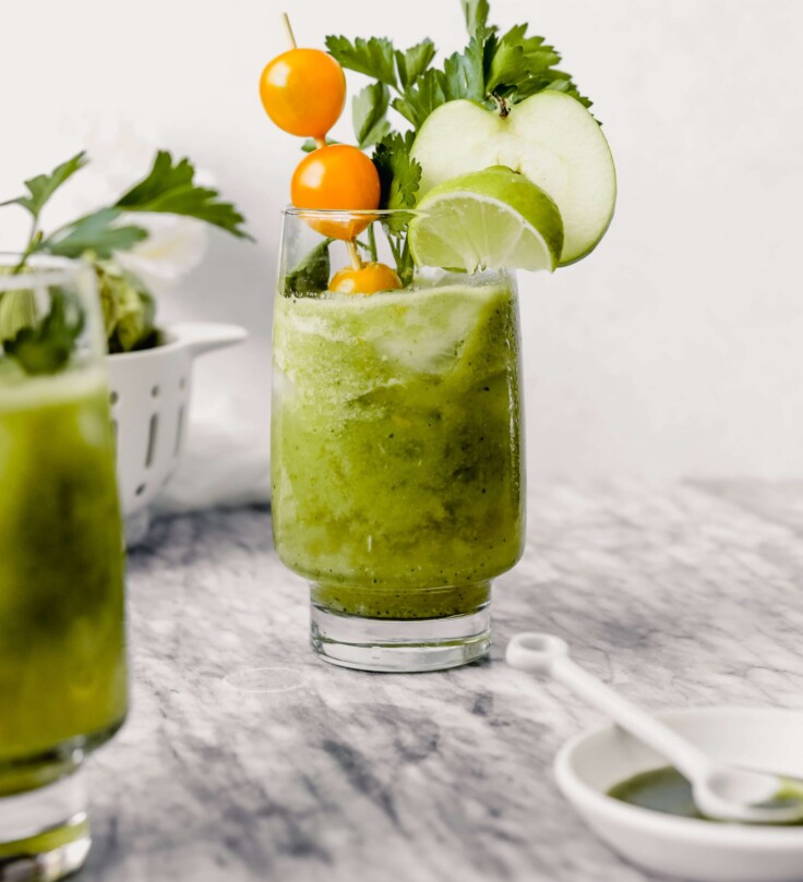Photograph of green bloody mary in tall collins glass garnishes with yellow cherry tomatoes, herbs, lime wedge and apple slice.