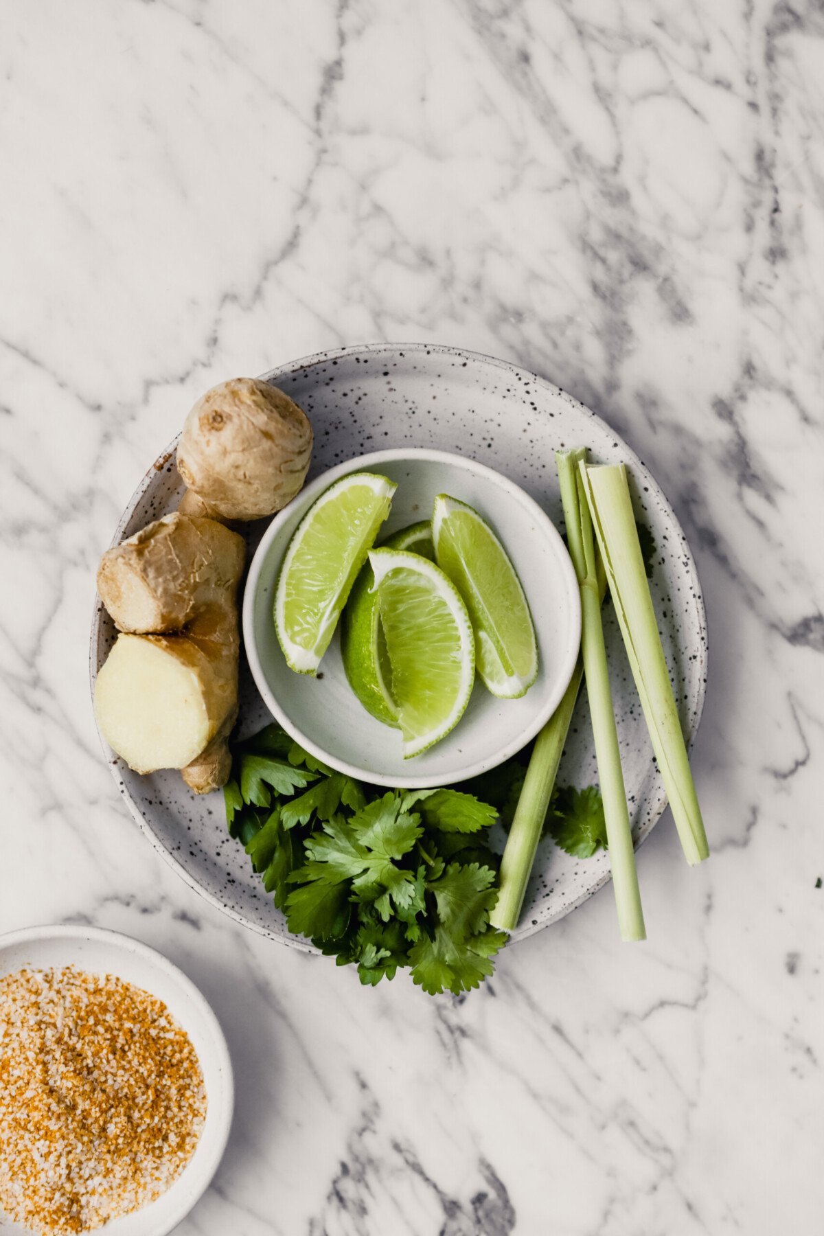 Photograph of ginger, lime wedges, lemongrass and fresh herbs arranged on a white speckled plate