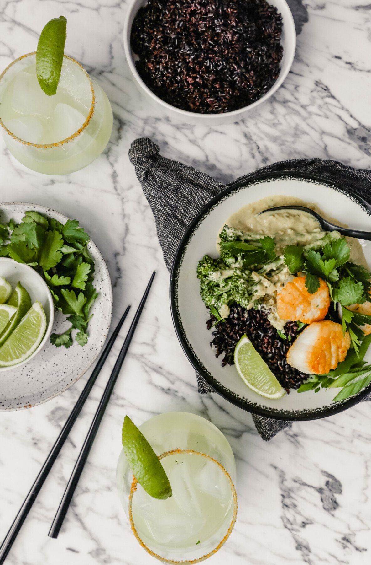 Photograph of a bowl filled with green coconut curry, black rice, broccolini and golden scallops set on a marble table with a gray napkin and margaritas set around.