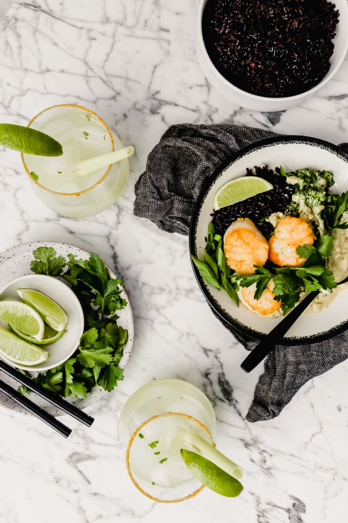 Photograph of a bowl filled with green coconut curry, black rice, broccolini and golden scallops set on a marble table with a gray napkin.