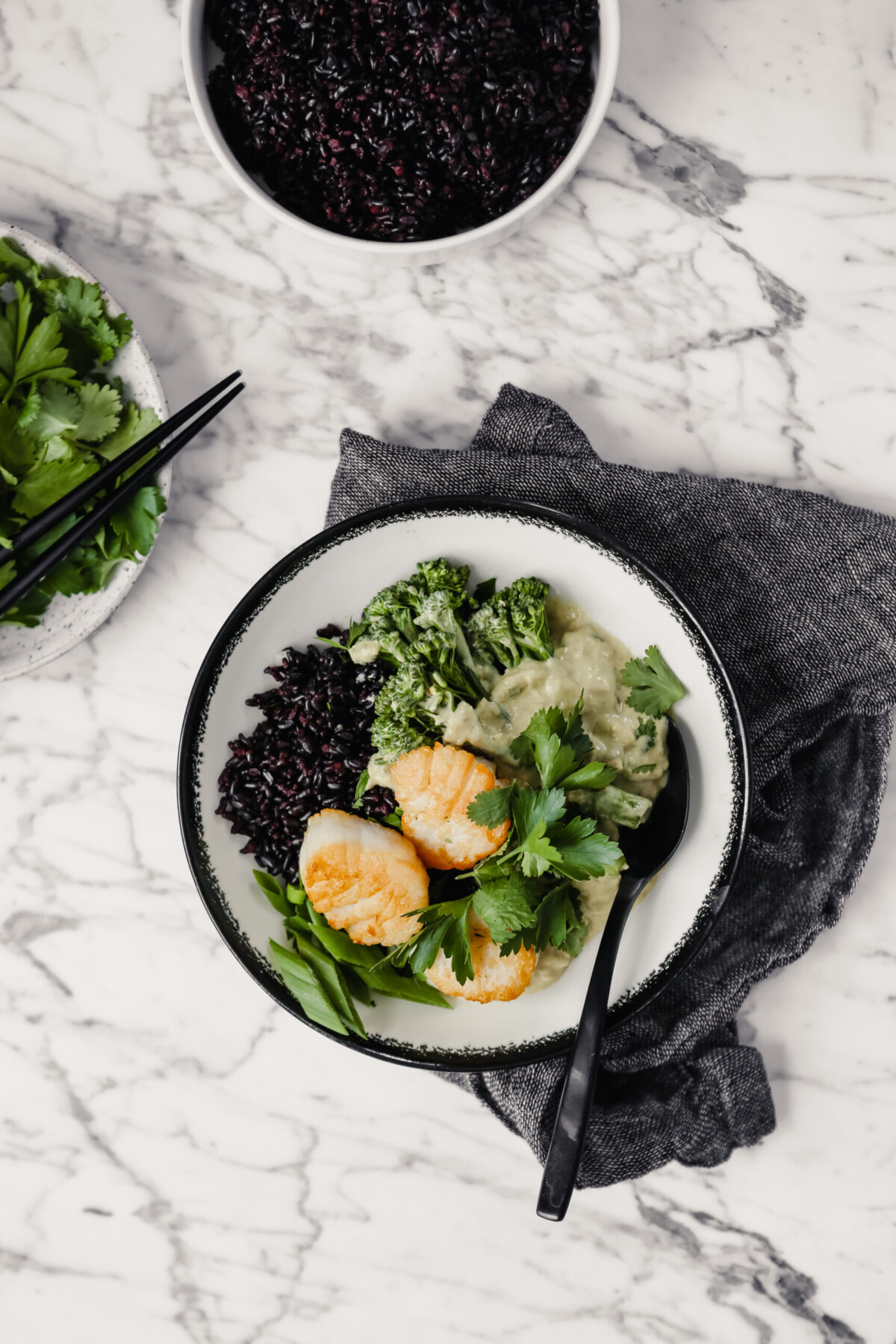 Photograph of a bowl filled with green coconut curry, black rice, broccolini and golden scallops set on a marble table with a gray napkin.