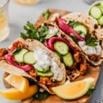 tacos filled with ground lamb, sliced cucumbers, pickled onions, herbs and a creamy sauce set on a wood cutting board with lemon wedges set around them