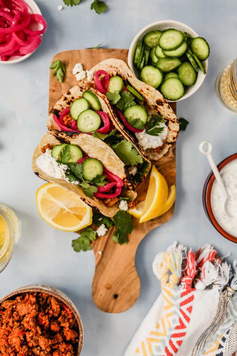 tacos filled with ground lamb, sliced cucumbers, pickled onions, herbs and a creamy sauce set on a wood cutting board with lemon wedges set around them
