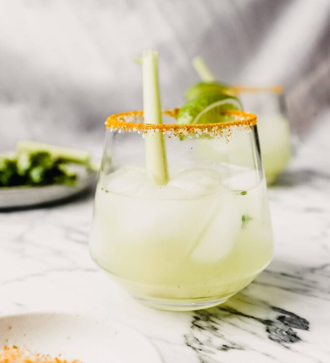Photograph of a margarita set on a white marble table garnished with a lime wedge and lemongrass.