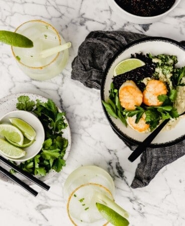 Photograph of a bowl filled with green coconut curry, black rice, broccolini and golden scallops set on a marble table with a gray napkin and margaritas set around.