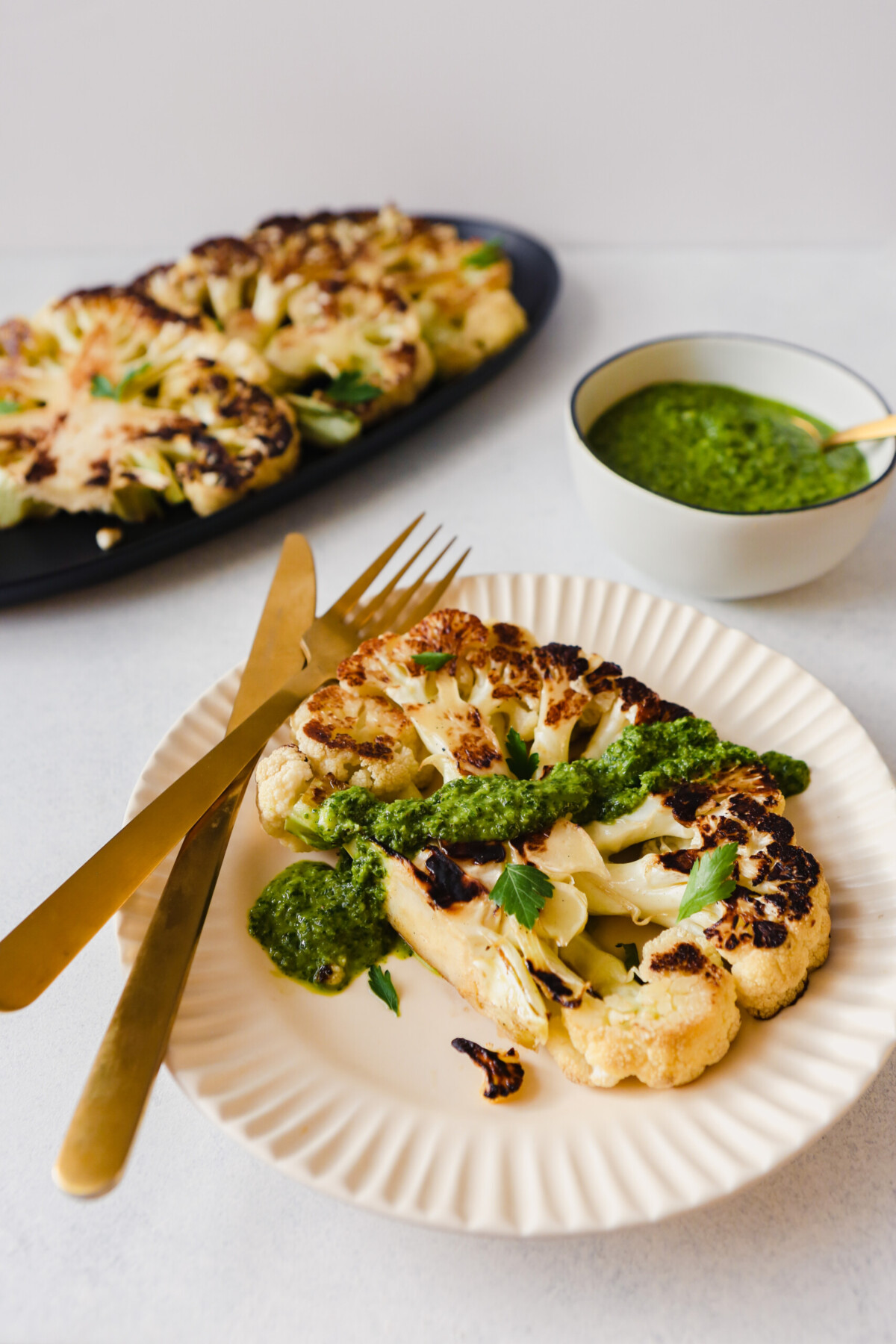 Photograph of cauliflower steaks set on a large serving platter, and once on a serving plate with a spread of green sauce over top