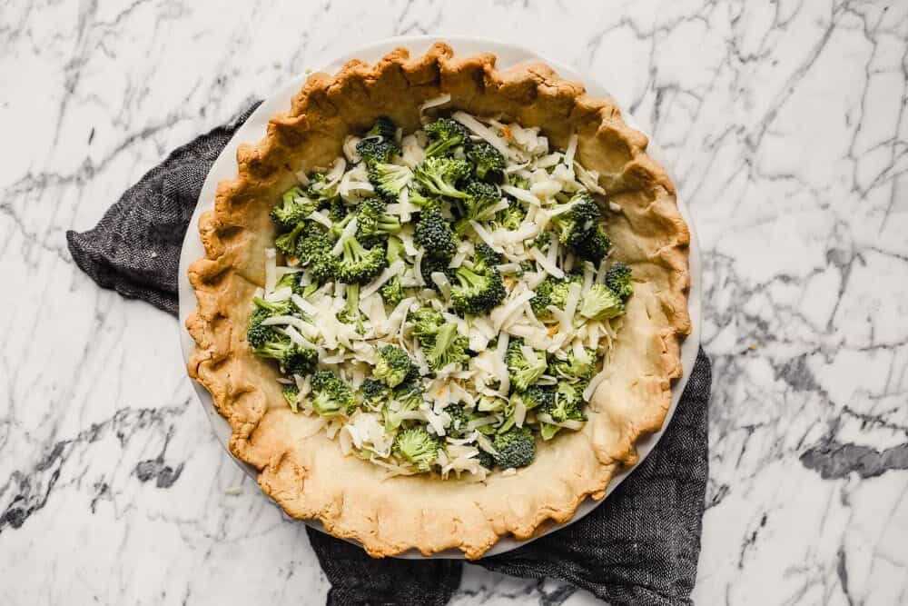 Photograph of cheese and broccoli sprinkled in the base of a baked pie shell