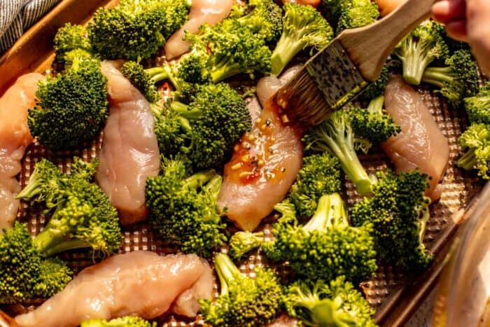 glaze being brushed on chicken tenders nestled on a baking sheet with broccoli