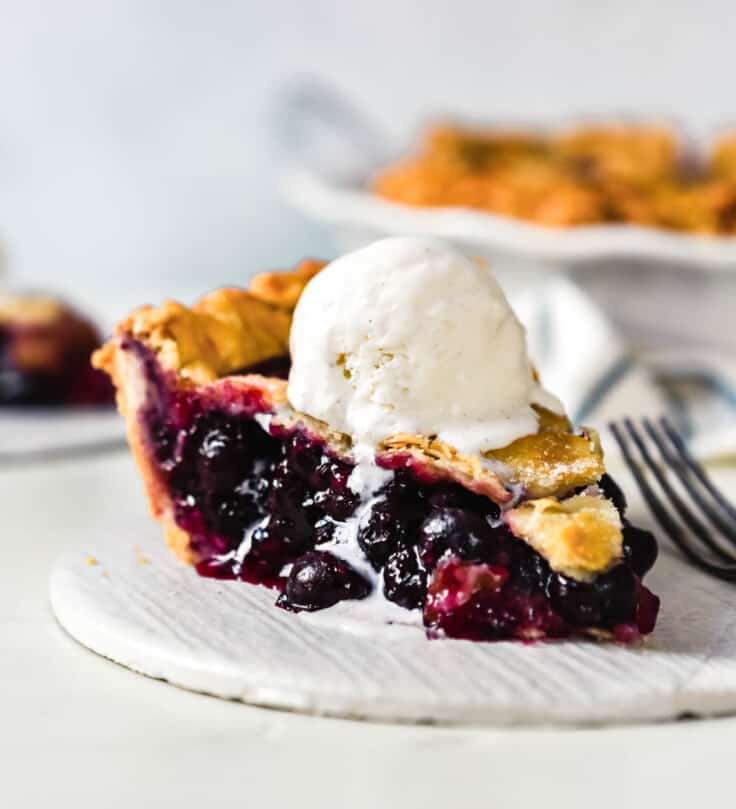 slice of blueberry pie set on a white plate with a scoop of vanilla ice cream on top and melting down the slice