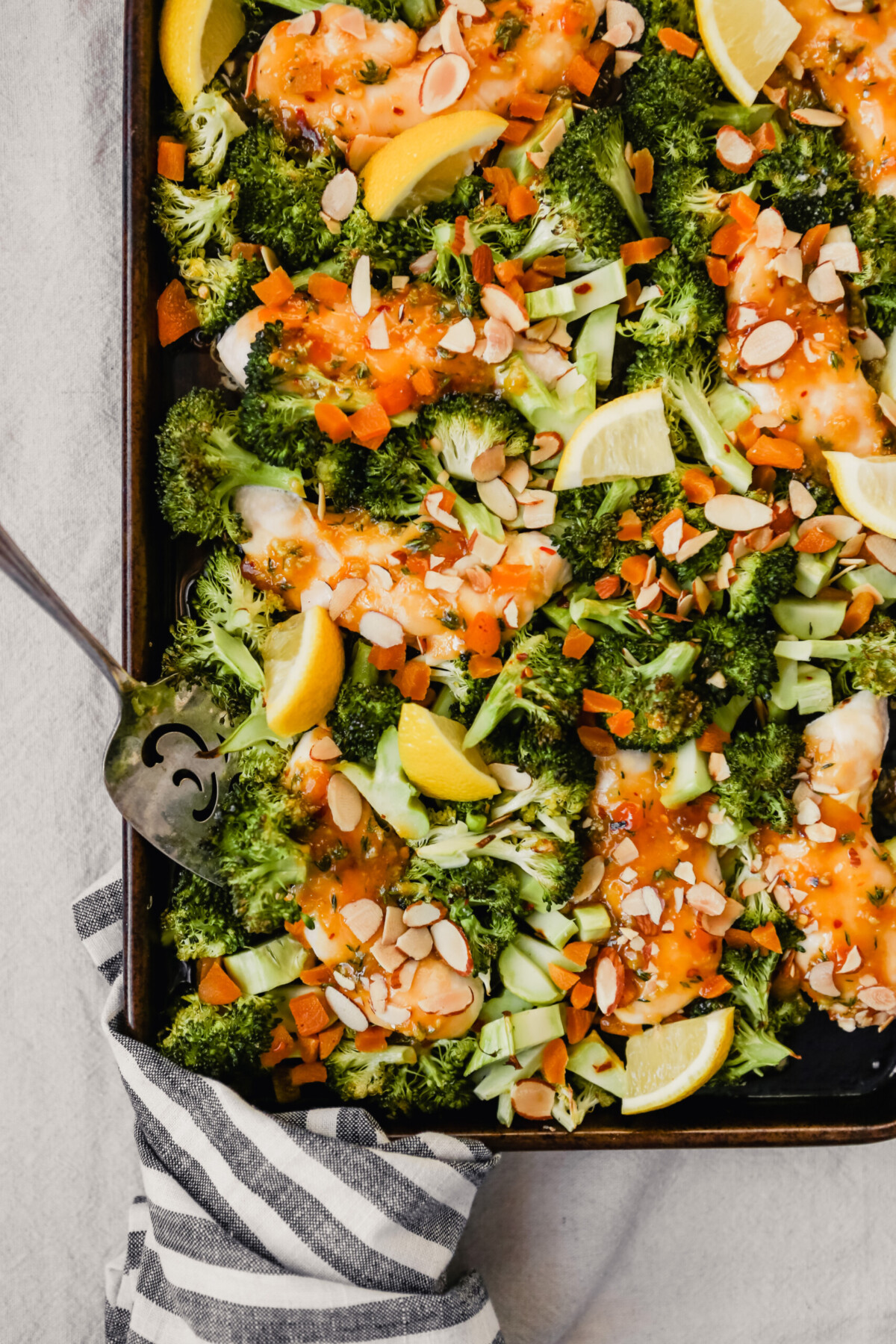 Photograph of apricot-glazed chicken on a sheet pan with broccoli, almond and lemon wedges.