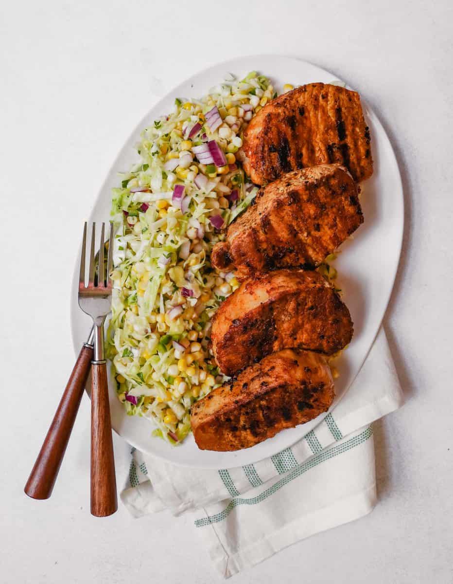 Photograph of grilled boneless pork chops on a white platter with sweet corn slaw.