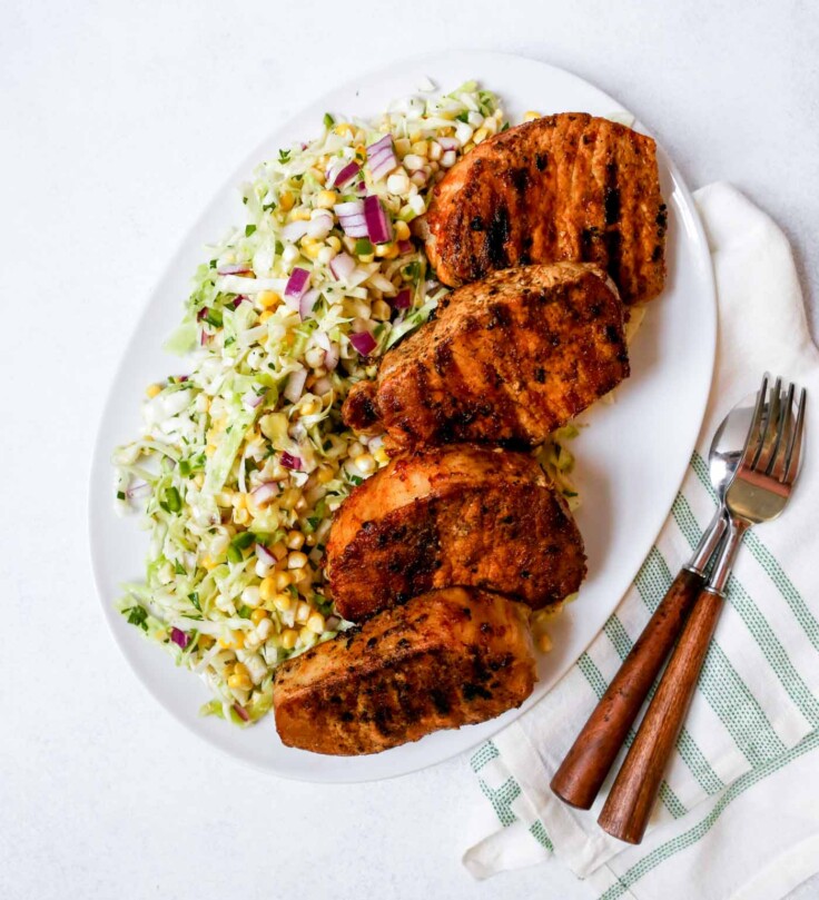 Photograph of grilled boneless pork chops on a white platter with sweet corn slaw.