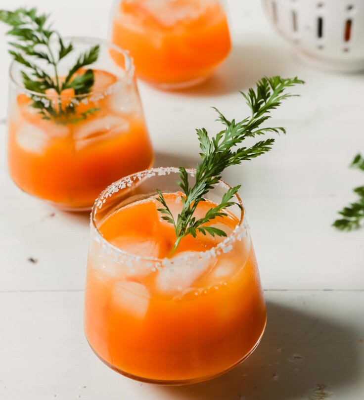 Photograph of carrot cocktails with carrot tops as garnish set on a white wood table.