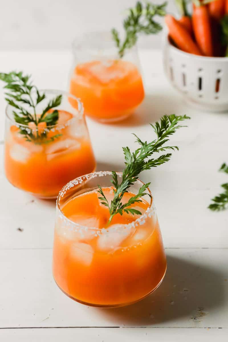 Photograph of carrot cocktails with carrot tops as garnish set on a white wood table.