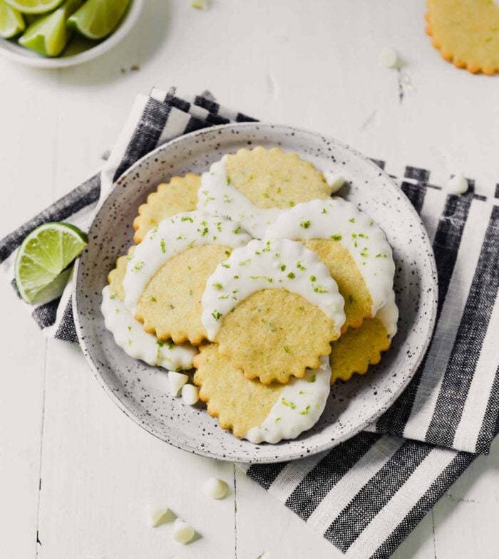 Photograph of glazed cornmeal cookies stacked on a speckled plate on a white table with limes scattered about
