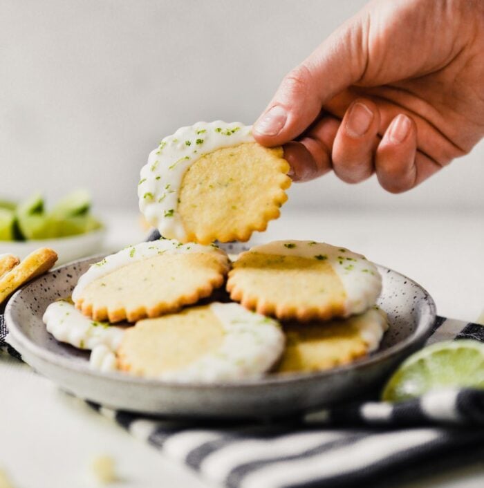 Photograph of glazed cornmeal cookies stacked on a speckled plate one being taken off by someone, set on a white table with limes scattered about