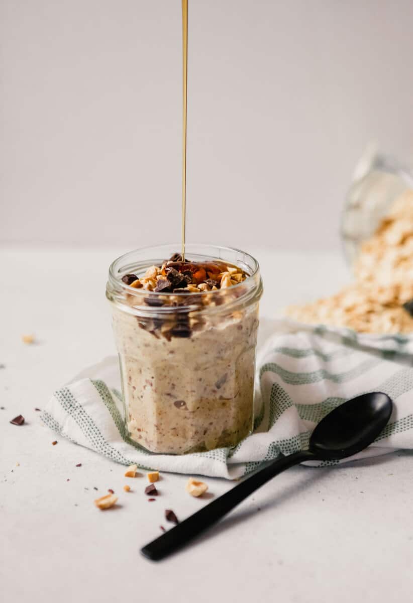 Photograph of chocolate peanut butter overnight oats in a glass jar on a table with spilled oats and maple syrup being poured over the oats