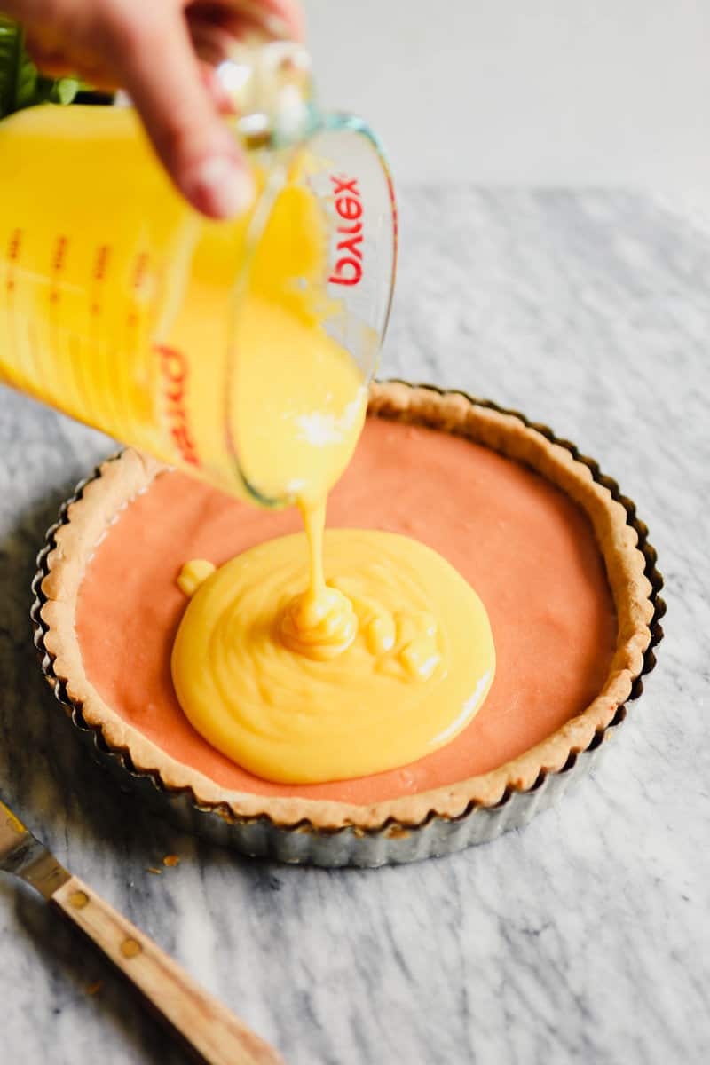 Photograph of a tart crust being filling with lemon curd set on a marble table.