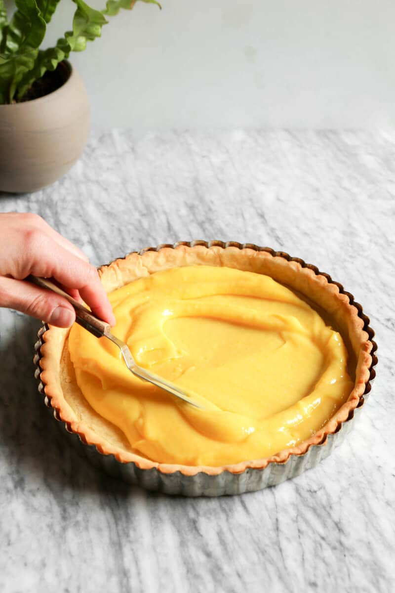 Photograph of a tart crust being filling with lemon curd set on a marble table.