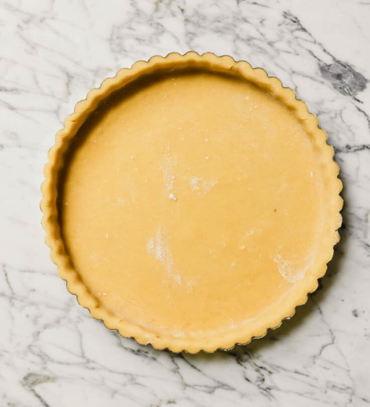 Photograph of an unbaked tart shell in a tart pan set on a marble table.