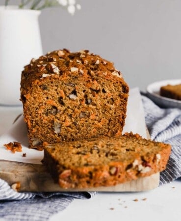 Photograph of carrot bread in a loaf pan set on a cutting board with a slice cut into it