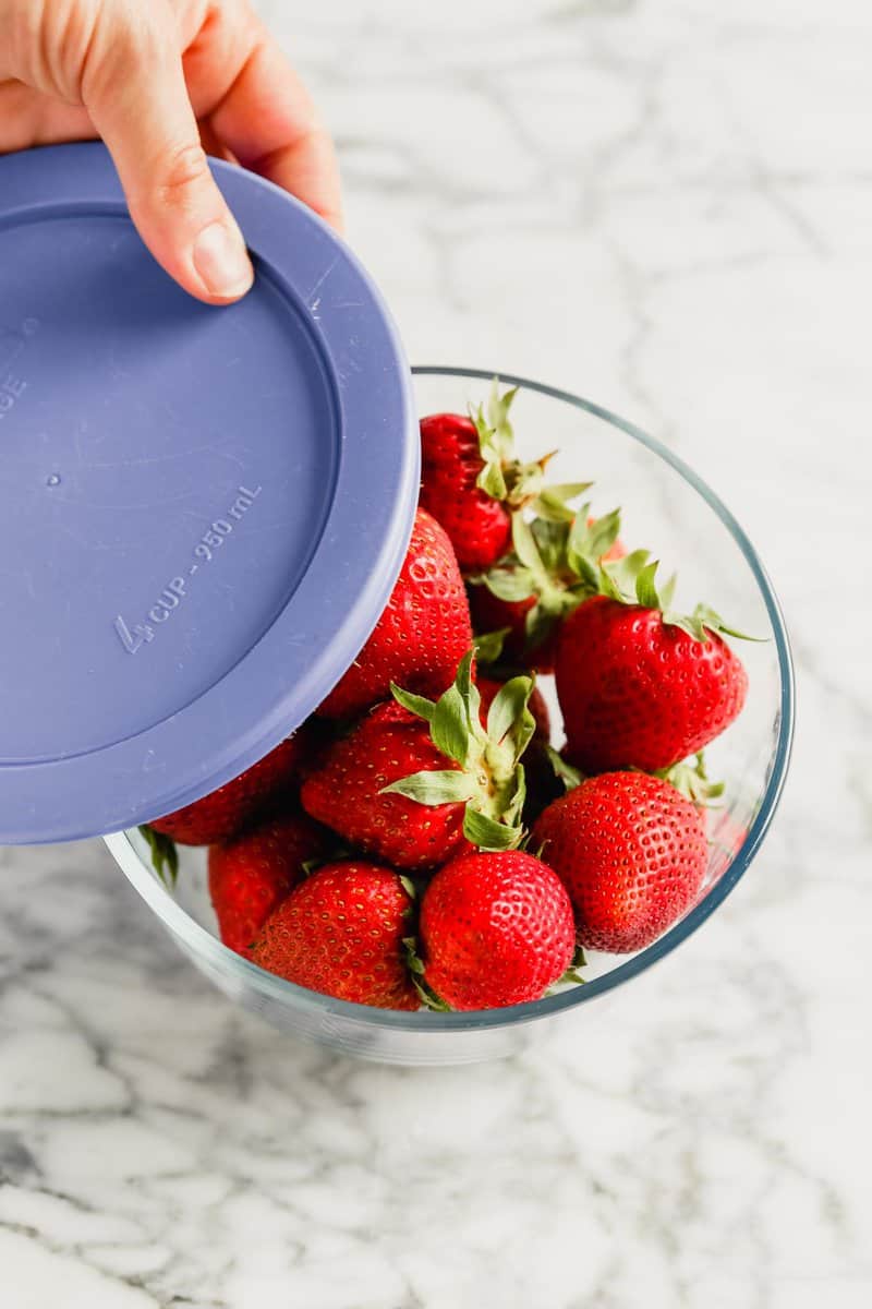 Photograph of fresh strawberries in a glass storage container with a blue lid set on a marble table