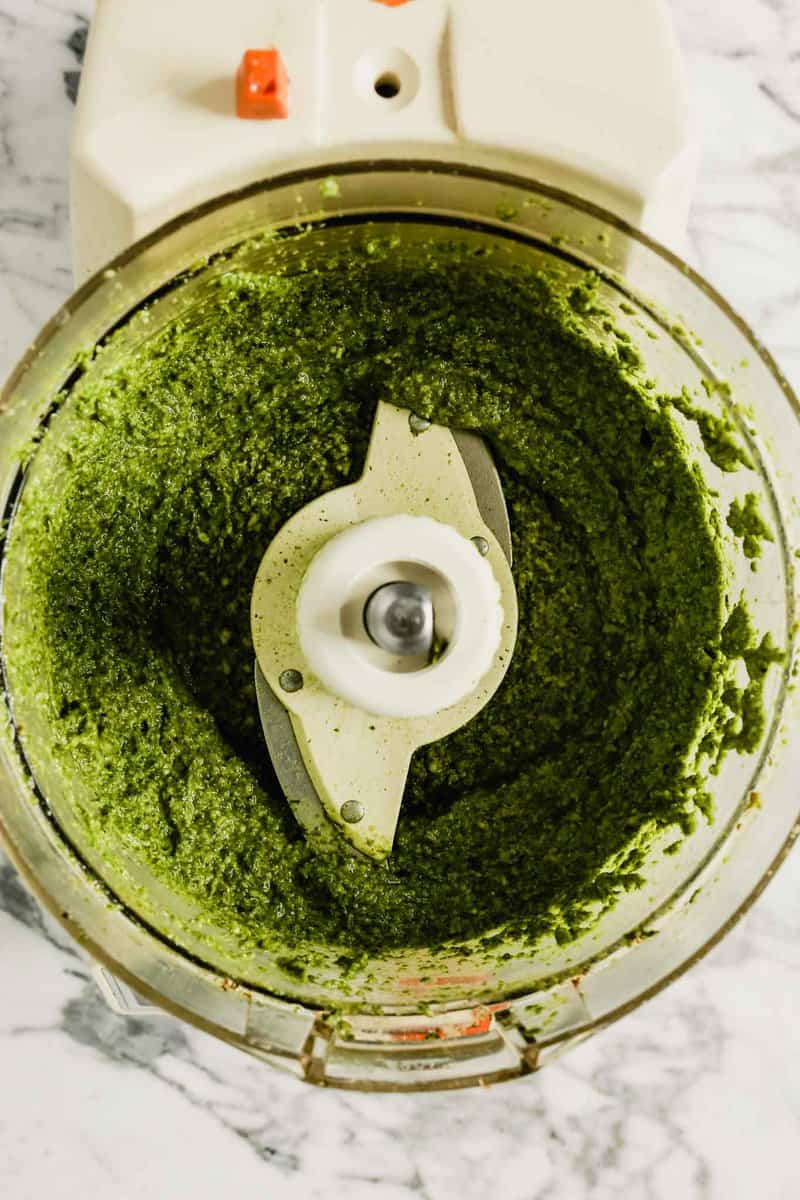 Photograph of bright green carrot top pesto in a food processor bowl set on a marble counter 