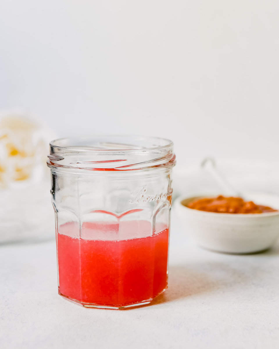 Photograph of pink-colored rhubarb simple syrup in a glass jar with sugar in the background
