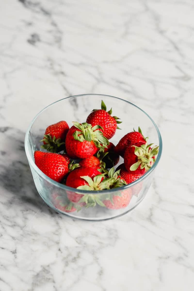 Photograph of fresh strawberries in a glass storage container set on a marble table