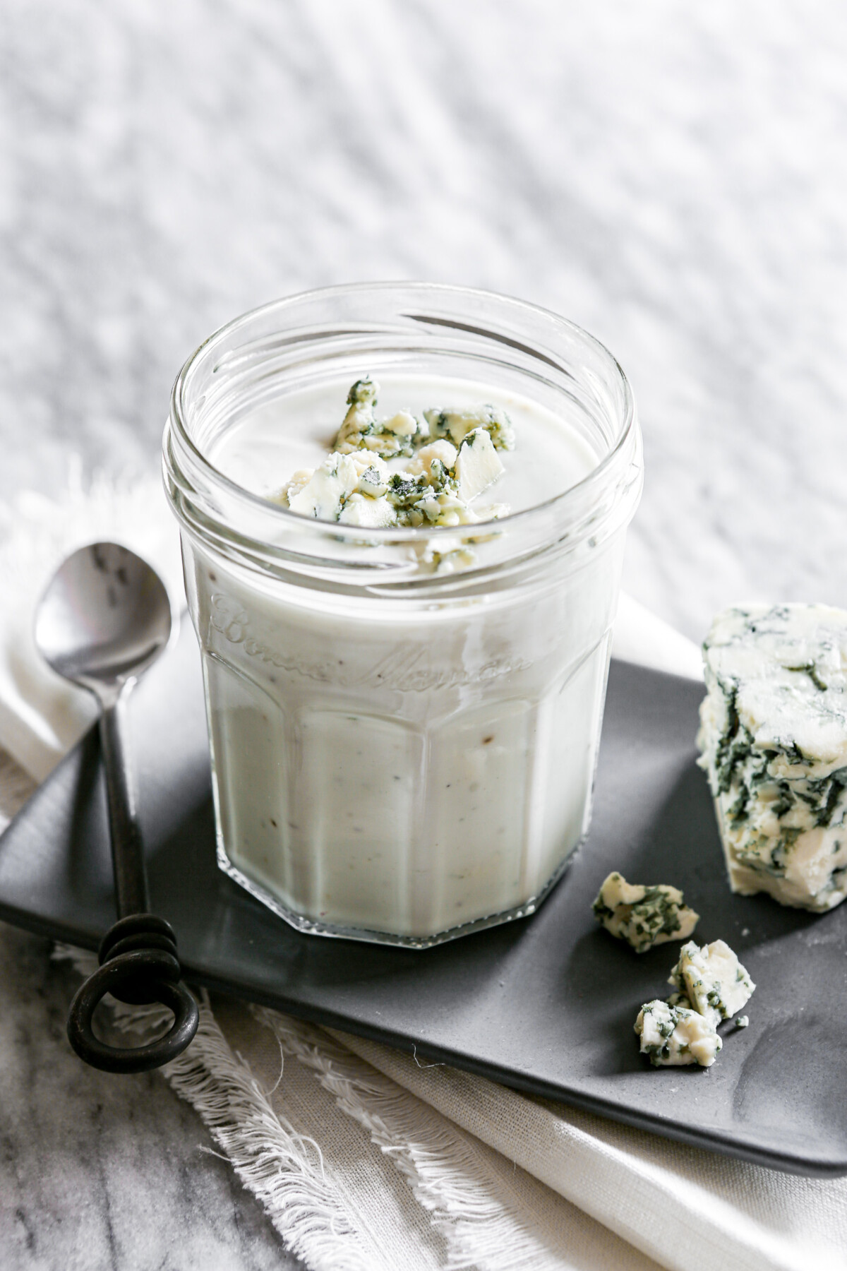 Photograph of blue cheese salad dressing in a glass jar set on a gray plate on a marble table.