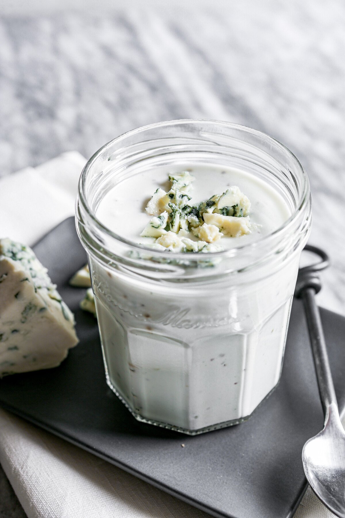 Photograph of blue cheese salad dressing in a glass jar set on a gray plate on a marble table.