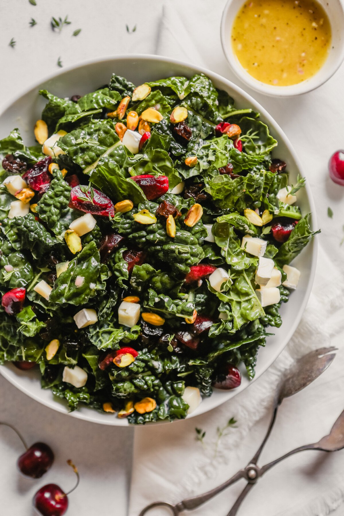 Photograph of a kale salad with cherries and pistachios in a white bowl on a blue table