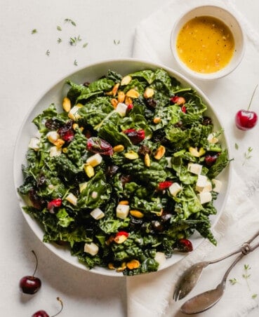 Photograph of a kale salad with cherries and pistachios in a white bowl on a blue table
