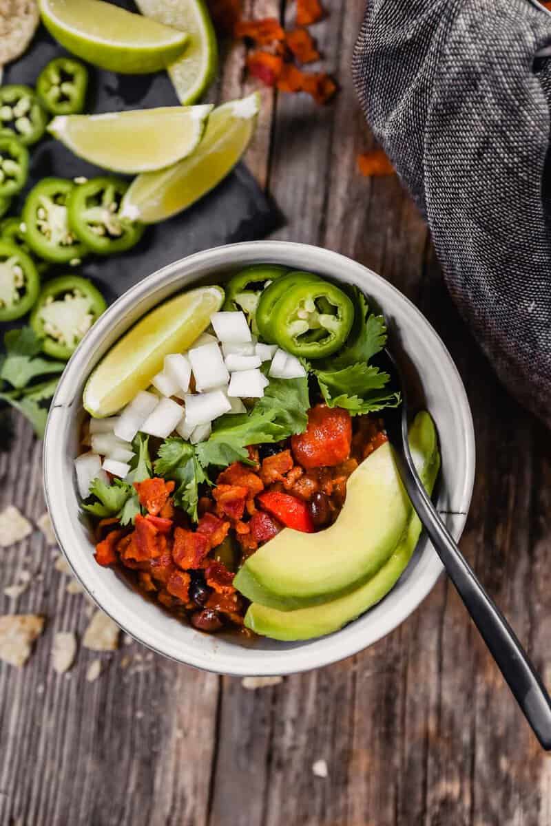 Photo of a bowl of chili topped with avocado, lime, onion, cilantro and jalapeno set on a wood table with chips