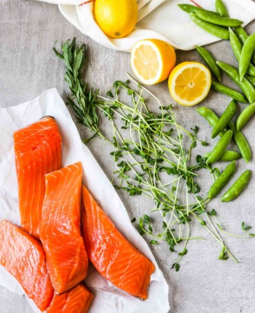 Overhead photo of fresh salmon filets stacked on a white paper with lemon, peas and herbs scattered around them.