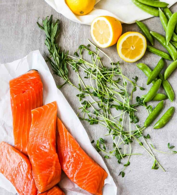 Overhead photo of fresh salmon filets stacked on a white paper with lemon, peas and herbs scattered around them.